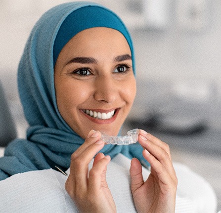 Woman in blue head covering smiling holding Invisalign in dental chair